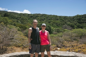 Stephan hiking Two people in casual outdoor attire stand side by side in front of a lush, green hillside under a clear blue sky.