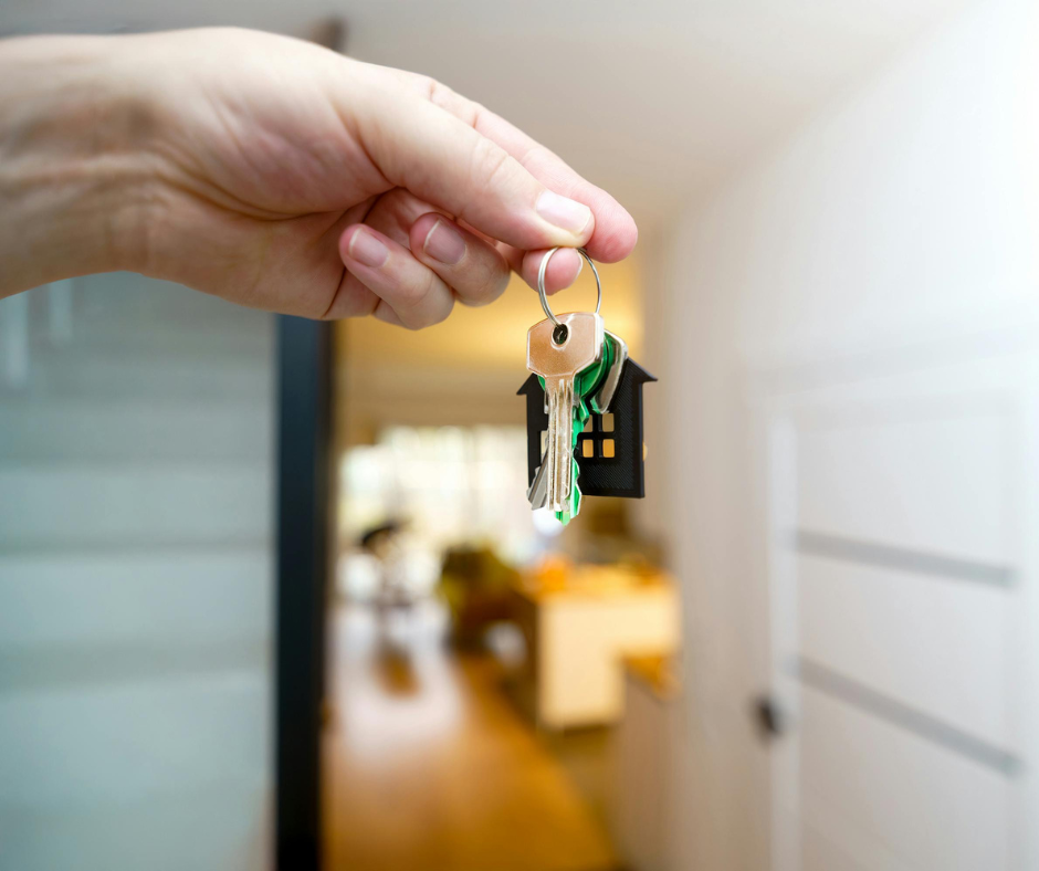 A hand holding a set of house keys with a black house-shaped keychain, in the foreground, with the interior of a house blurred in the background.